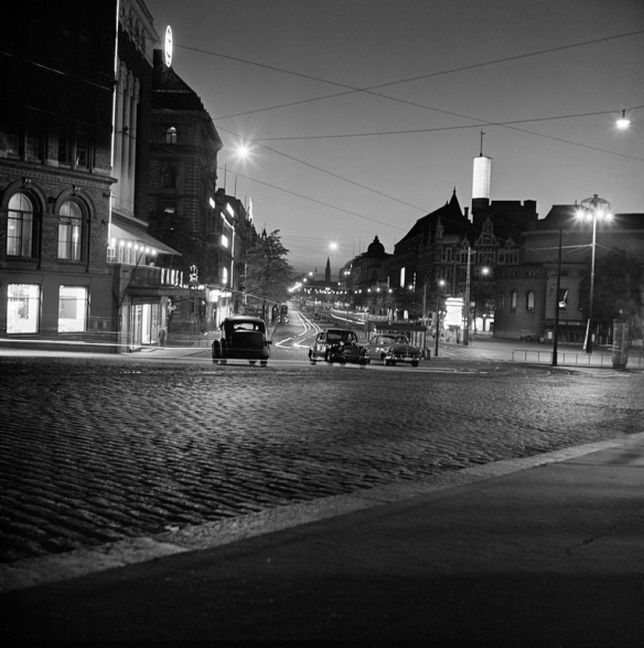 1953 . Liikennettä ja valoja öisellä Mannerheimintiellä. Kuvattu Erottajalta luoteeseen, vas. edessä Rake Oy (Erottajankatu 4), oik. edessä Ruotsalaisen teatterin kaareva seinä, sen takana Stokmann. Taustalla kesk. näkyy Kansallismuseon torni.